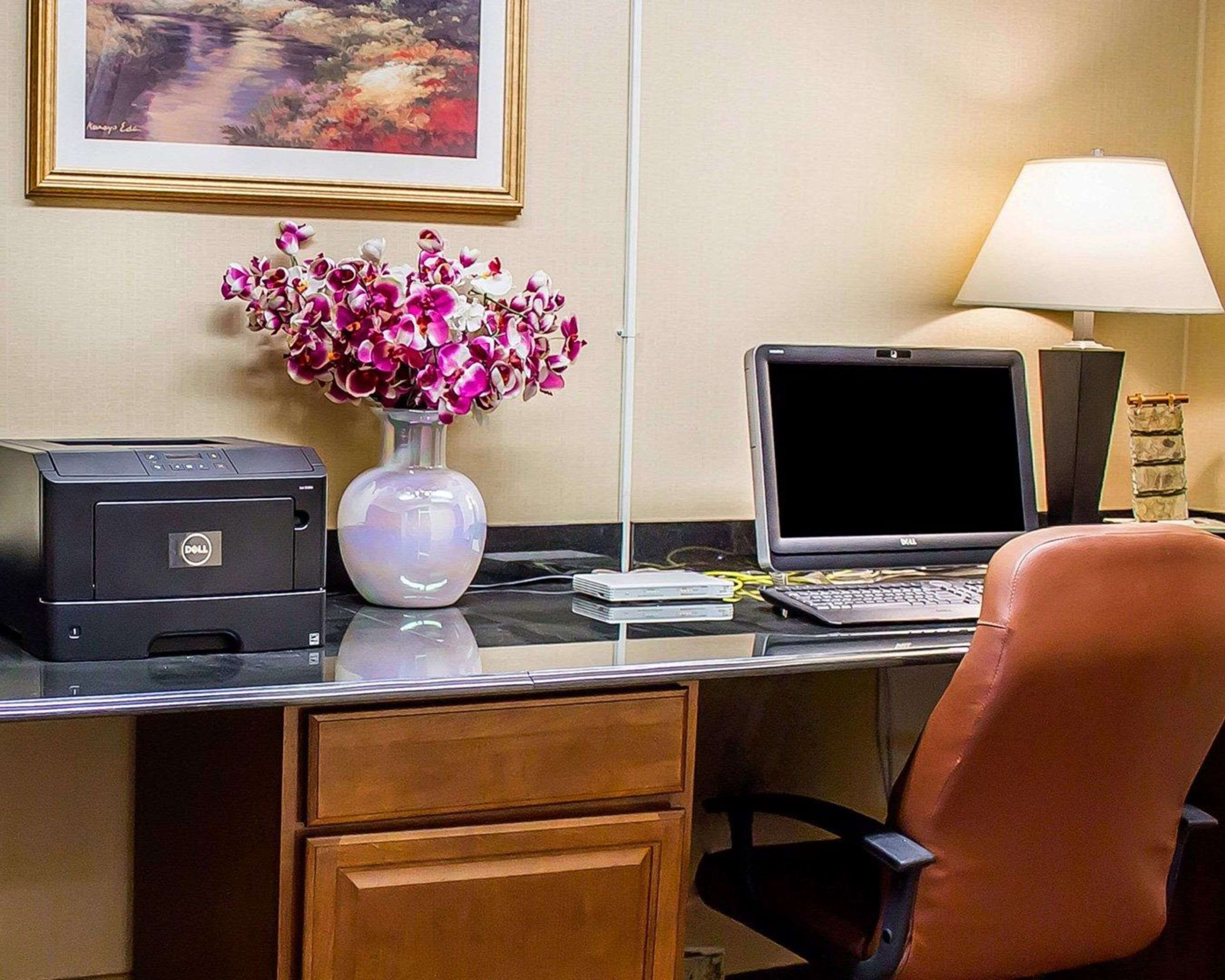 A hotel room desk with a Dell printer, a laptop, a vase of flowers, a lamp, and a framed picture on the wall.