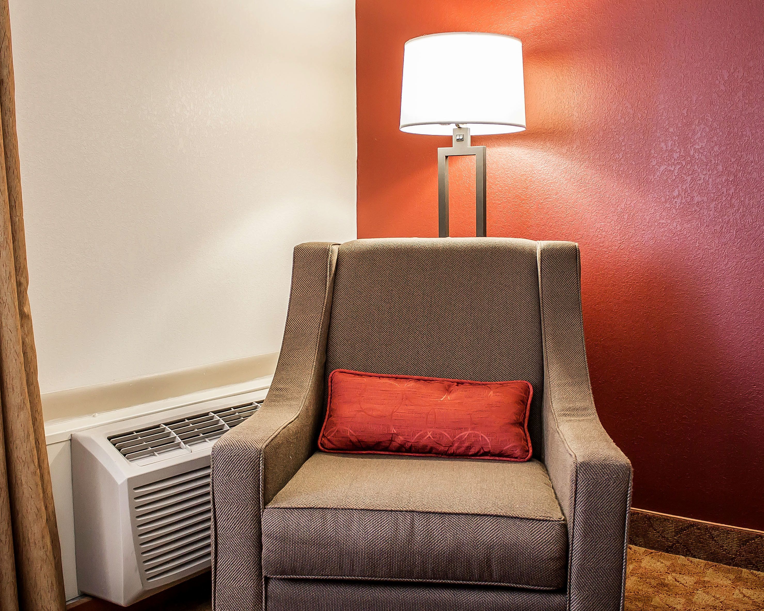 A chair with a herringbone pattern upholstery and a red cushion, positioned next to a wall heater and a floor lamp with a white lampshade.