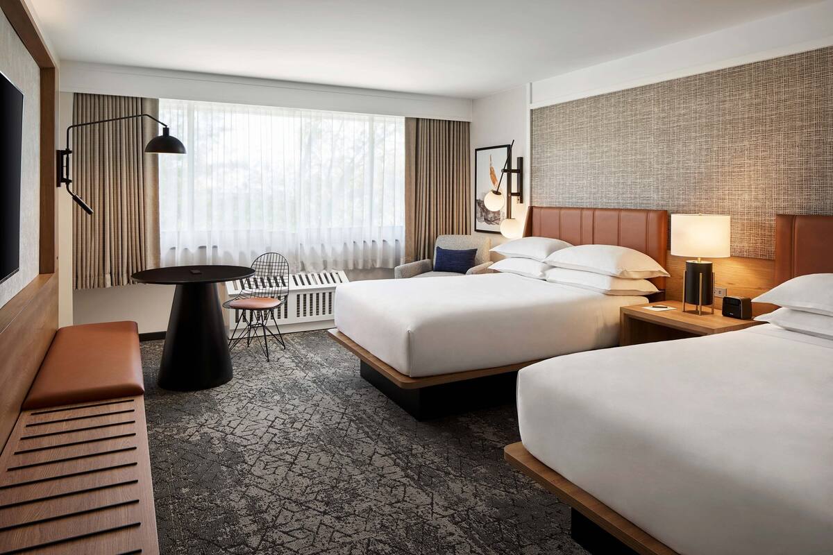 A hotel room with two beds, a desk, a chair, a TV, and a large window with curtains.