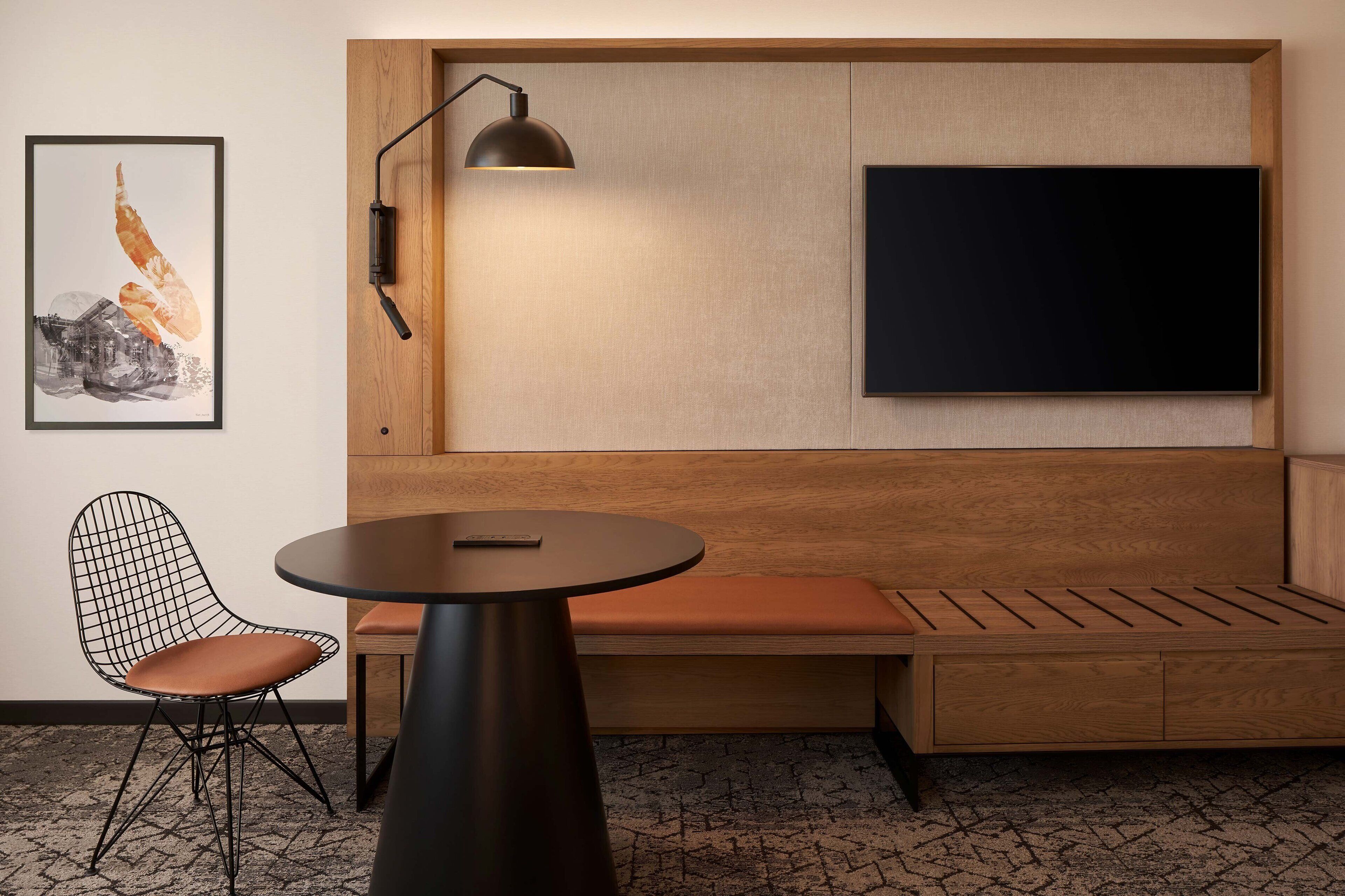 Una habitación moderna con un banco de madera, una mesa redonda, una silla negra, un televisor montado y un cuadro en la pared.