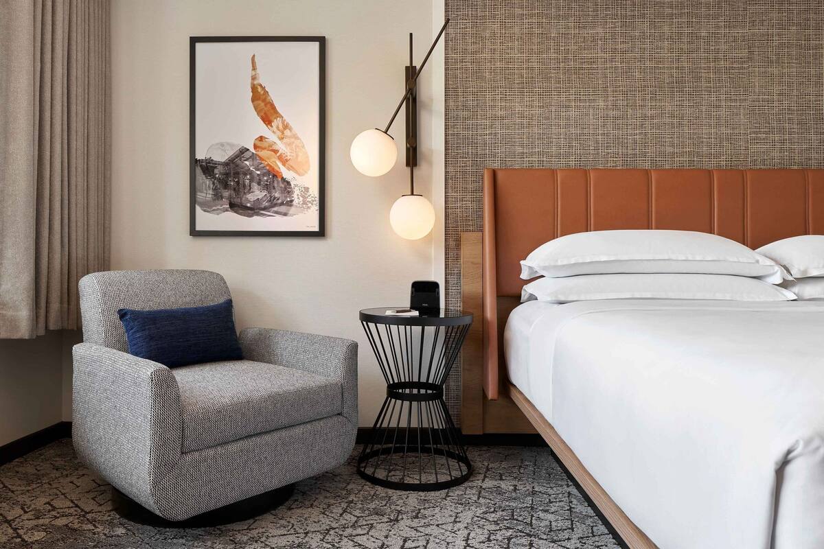 A hotel room with a bed, a chair, a small side table, a wall-mounted light fixture, and a framed picture on the wall.