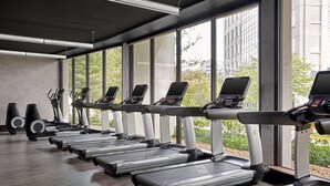 A gym with treadmills and elliptical machines arranged in a row, large windows, and a view of greenery and buildings outside.