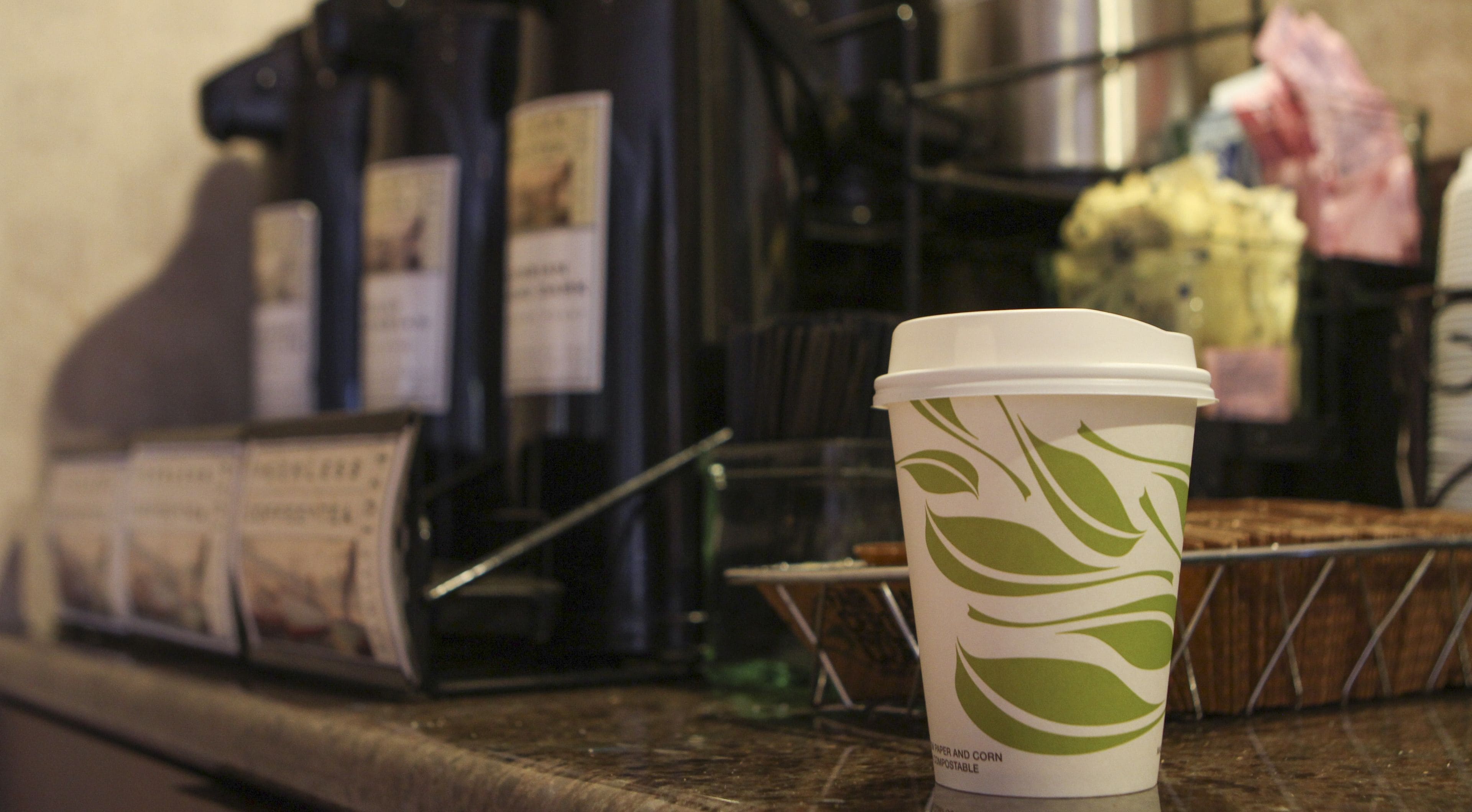 A coffee cup with a leaf design on a countertop, a coffee machine, and a muffin tray in the background.