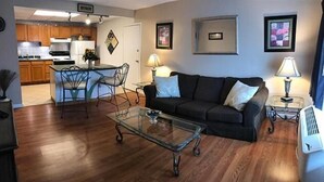 A living room with a glass coffee table, a dark sofa, and a dining area with wooden cabinets and a white refrigerator. - Wingfield Inn (Mayfield)