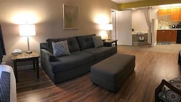 A living room with a dark grey sofa, two patterned throw pillows, a side table with a lamp, and a matching ottoman. A kitchen is visible in the background.