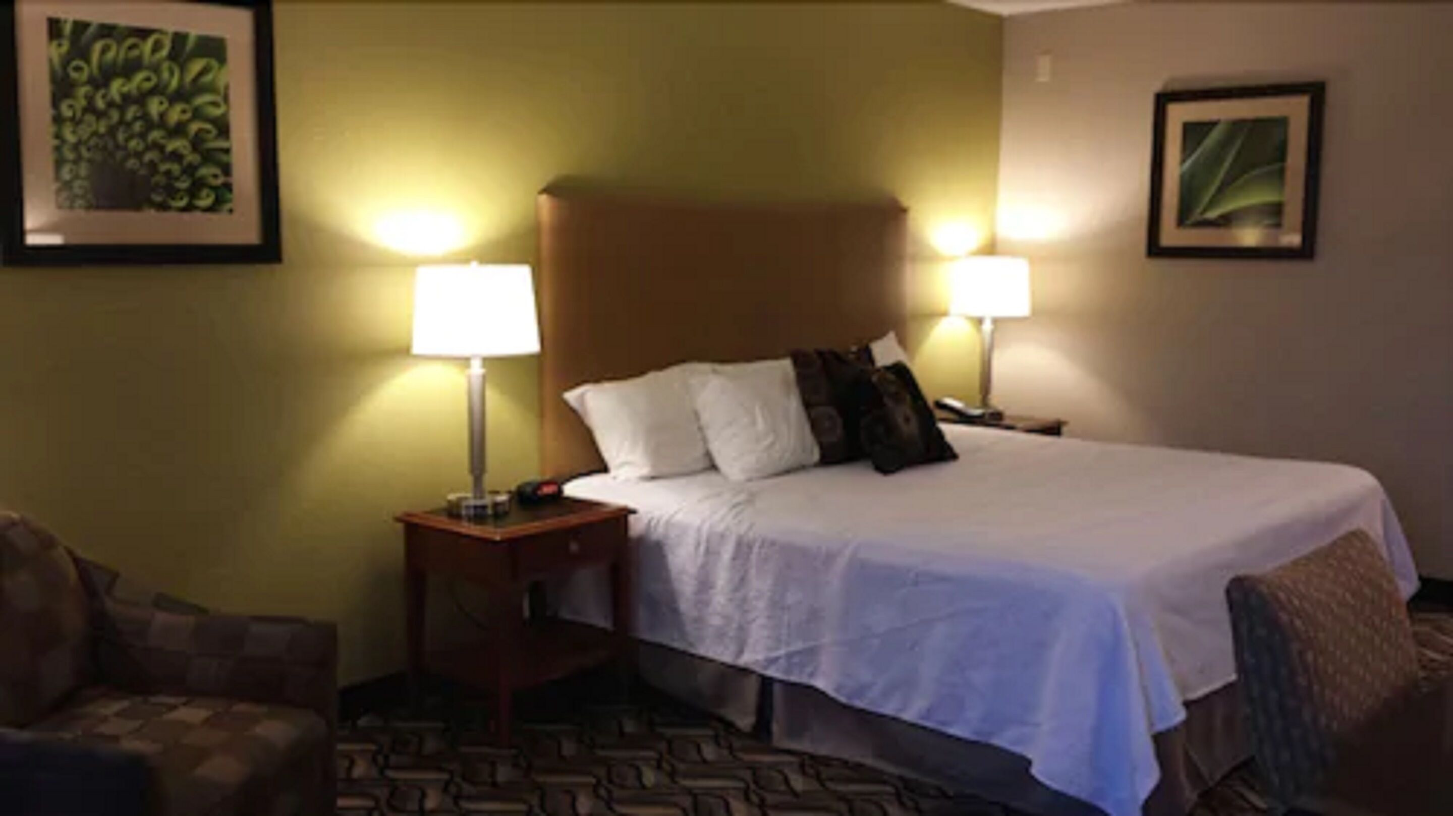 A hotel room with a bed, two bedside lamps, a headboard, a nightstand, a chair, and a patterned carpet.