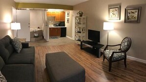 A living room with a sofa, ottoman, TV stand, and chair. There is a kitchen in the background. - Wingfield Inn (Mayfield)