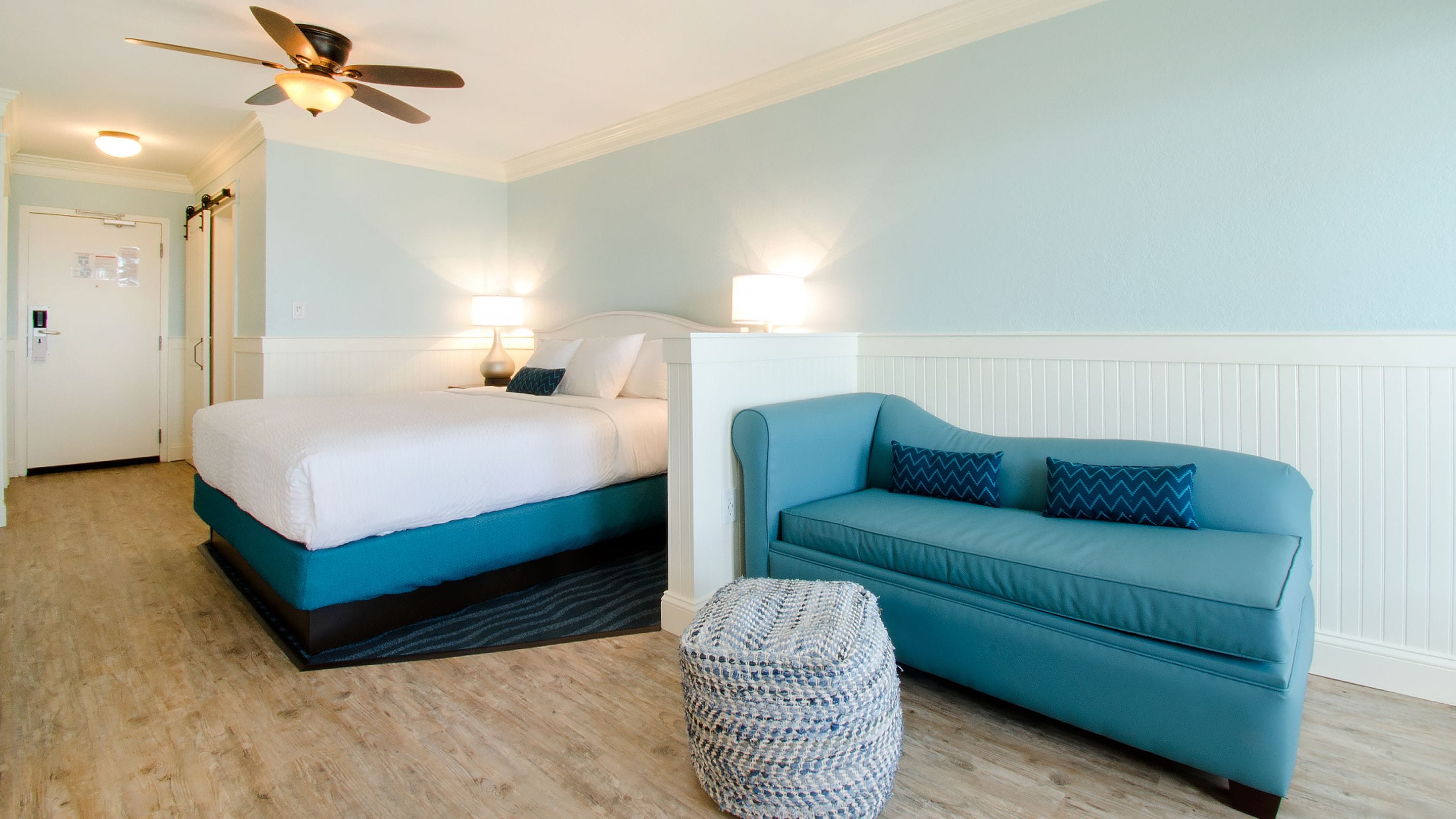 A hotel room with a bed, a blue sofa, a woven ottoman, a ceiling fan, and a wall-mounted lamp.