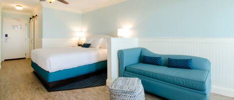 A hotel room with a bed, a blue sofa, a woven ottoman, a ceiling fan, and a wall-mounted lamp.