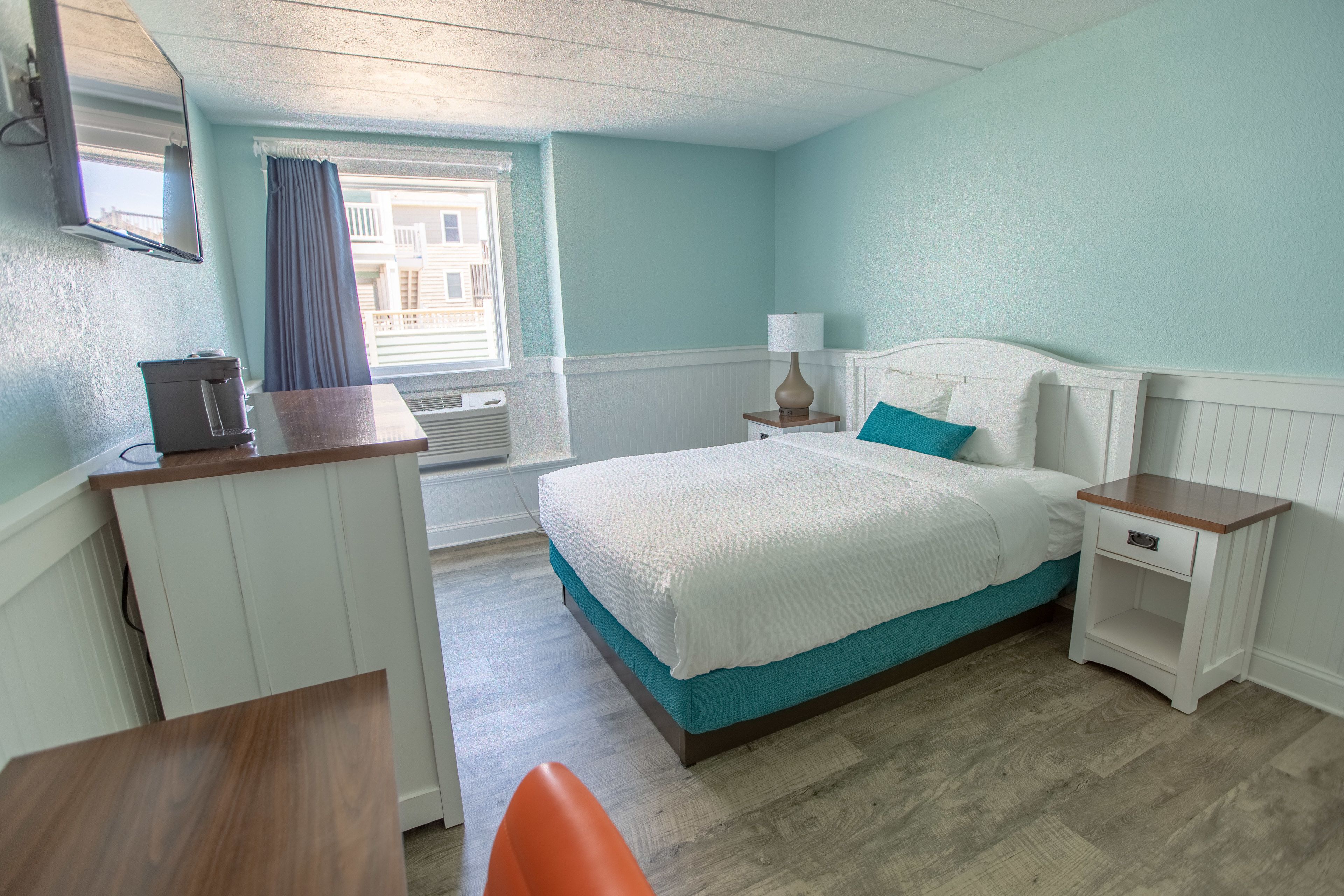 A bedroom with a bed, a desk, a chair, a nightstand, a lamp, and a window with curtains.