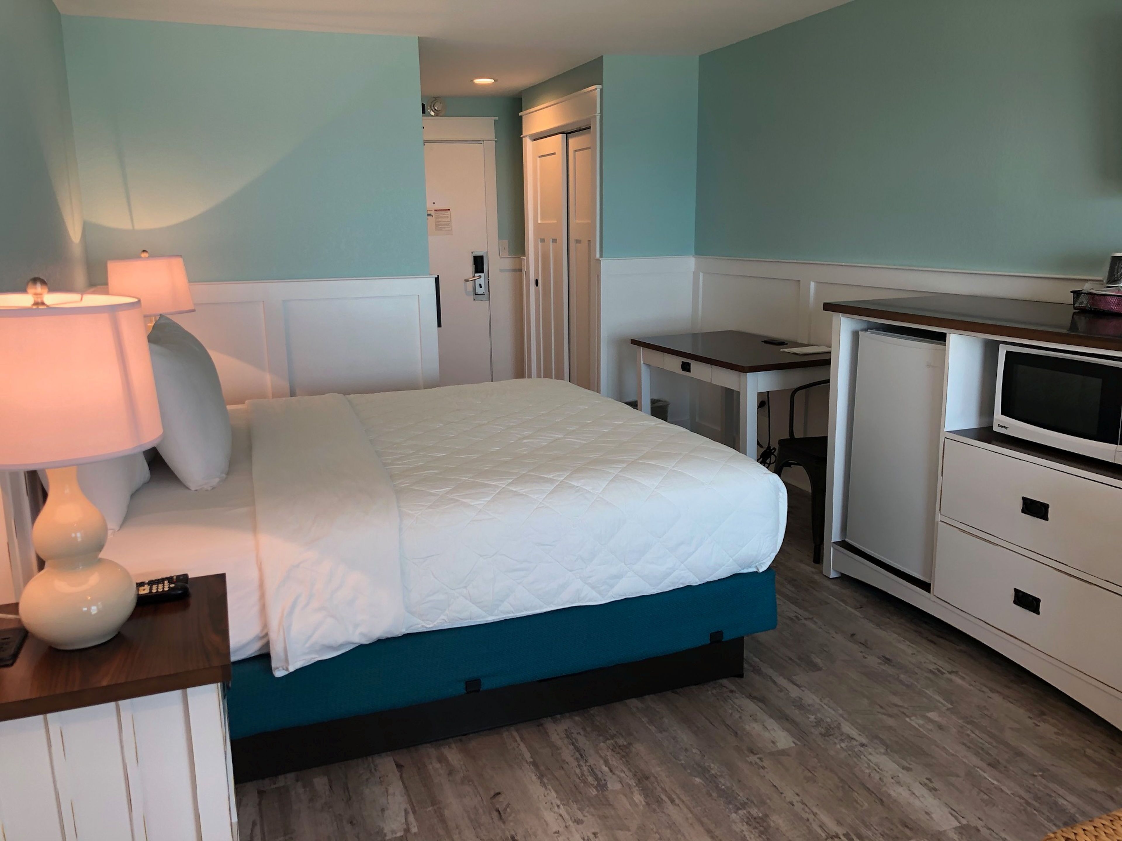 A bedroom with a bed, bedside lamp, dresser, TV, and a door leading to another room.