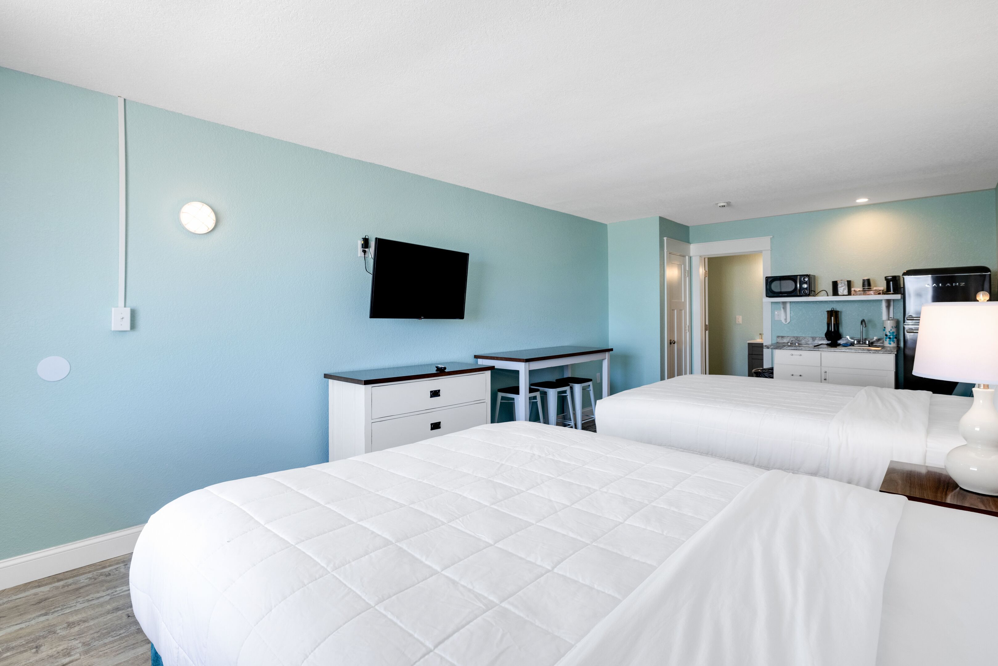 A hotel room with two beds, a desk, a TV, and a kitchenette.