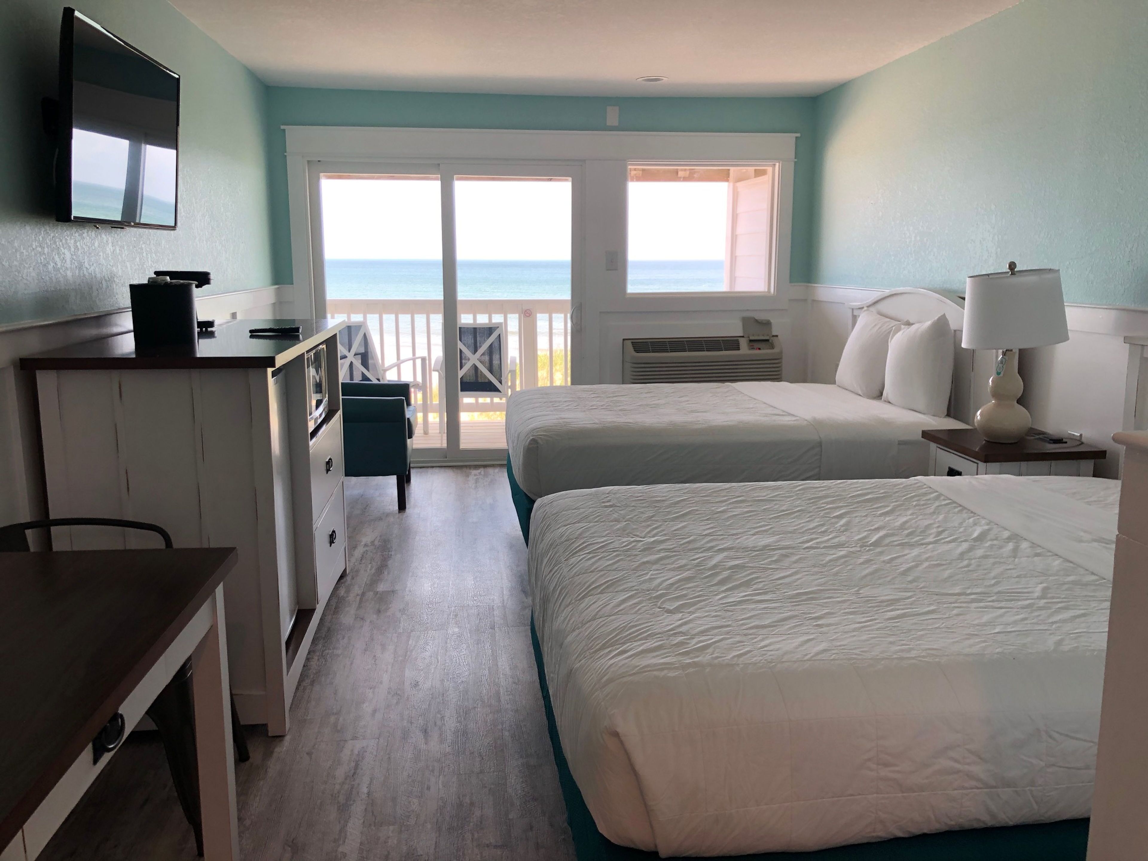 A hotel room with two beds, a desk, a TV, and a balcony with a view of the ocean.