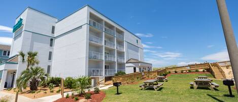 A multi-story hotel with a white exterior, a grassy lawn, picnic tables, and a wooden staircase leading to the entrance.