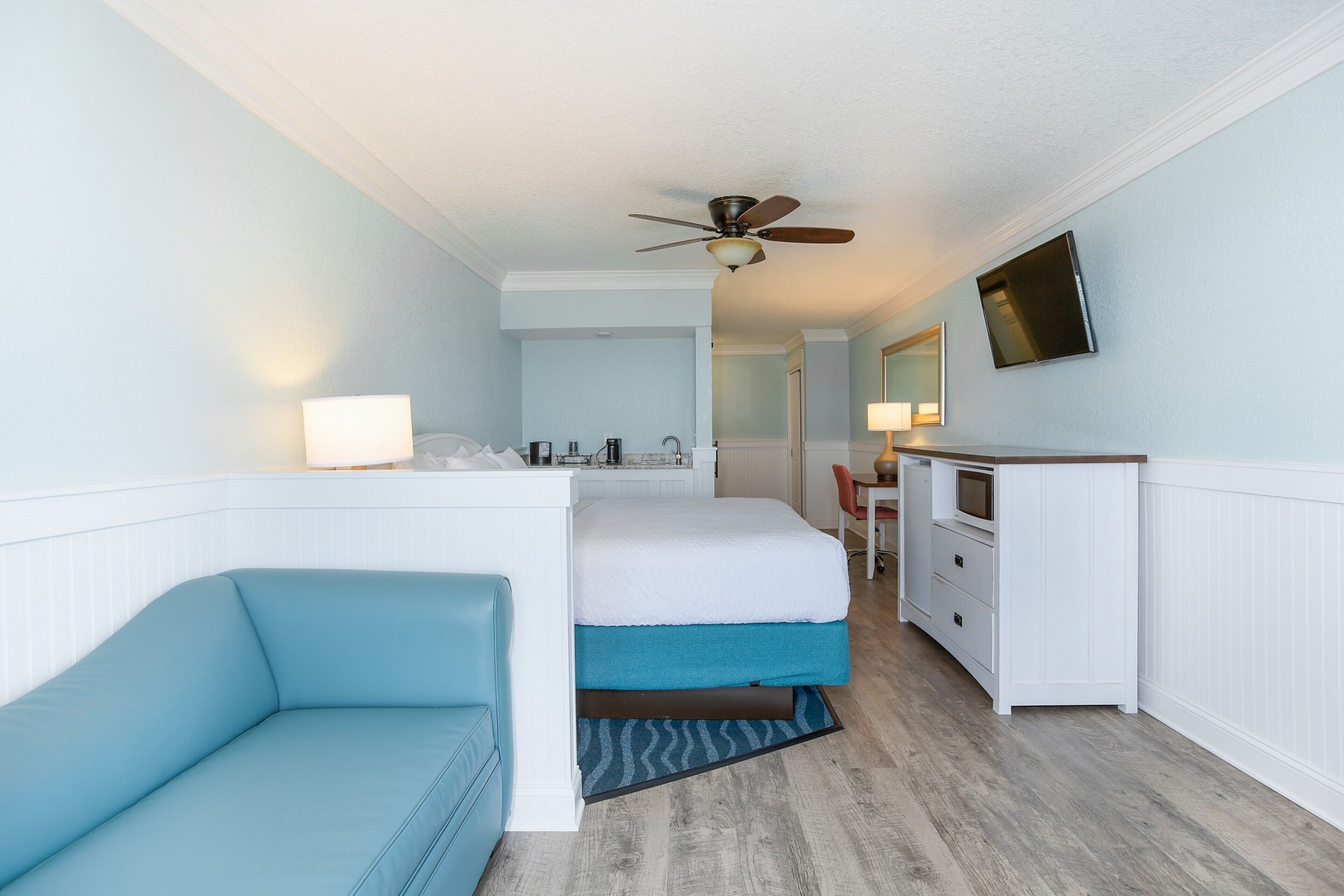 A hotel room with a bed, a blue sofa, a desk with a chair, a television, and a ceiling fan.