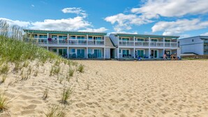 Un hotel frente a la playa con varios balcones, una playa arenosa al frente y cielos azules despejados.