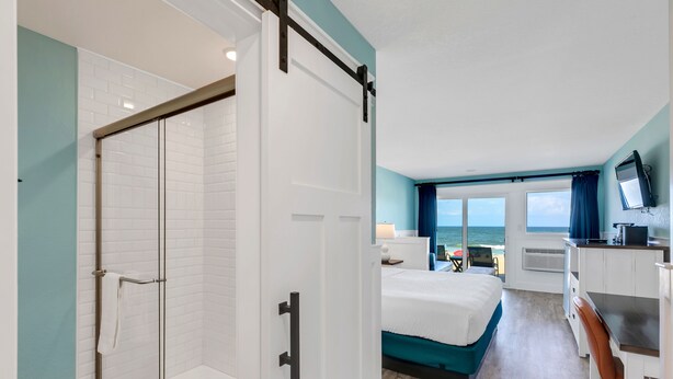 Habitación de hotel con ducha de vidrio, cama con cabecera azul, escritorio con computadora y vista al océano a través de ventanas amplias.