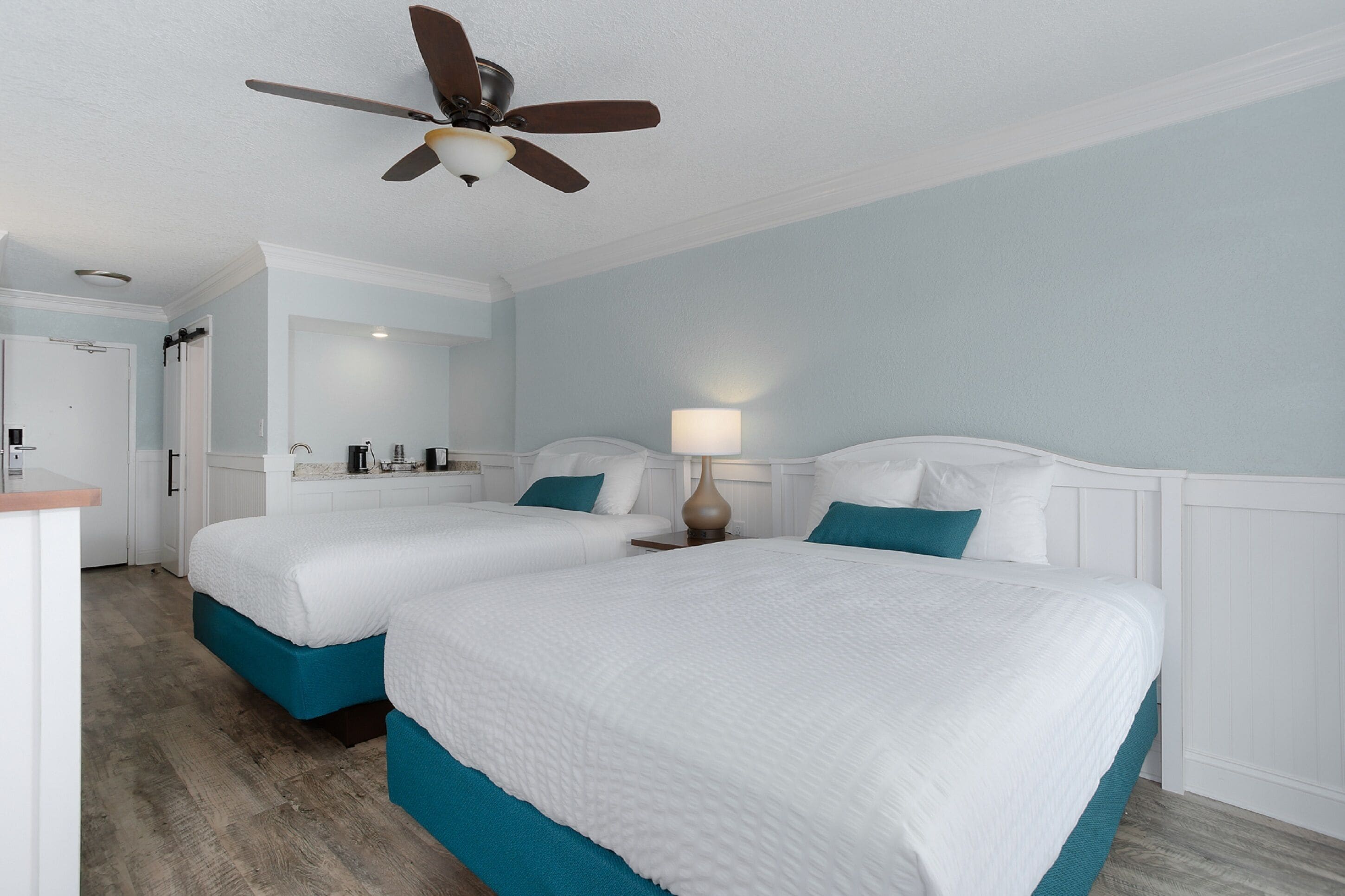 Une chambre d’hôtel avec deux lits, un ventilateur de plafond, une salle de bain avec douche et une kitchenette.