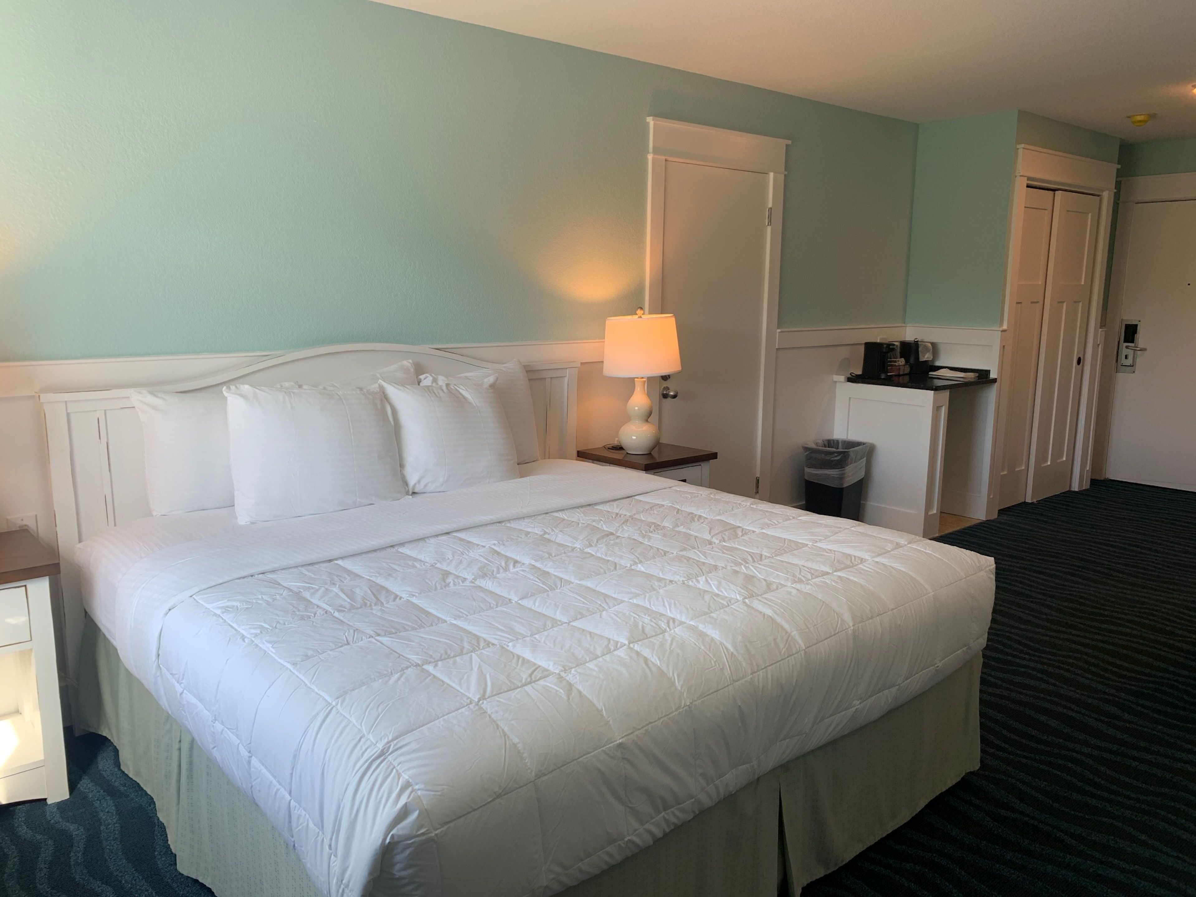 A hotel room with a large bed, a bedside lamp, a small table, a trash bin, and a wardrobe.