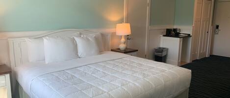 Une chambre d’hôtel avec un grand lit, une lampe de chevet, une petite table, une poubelle et une armoire.