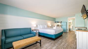 A modern hotel room with a teal sofa, a wooden bench, a bed with white bedding, a small kitchenette, and a flat-screen TV mounted on the wall.