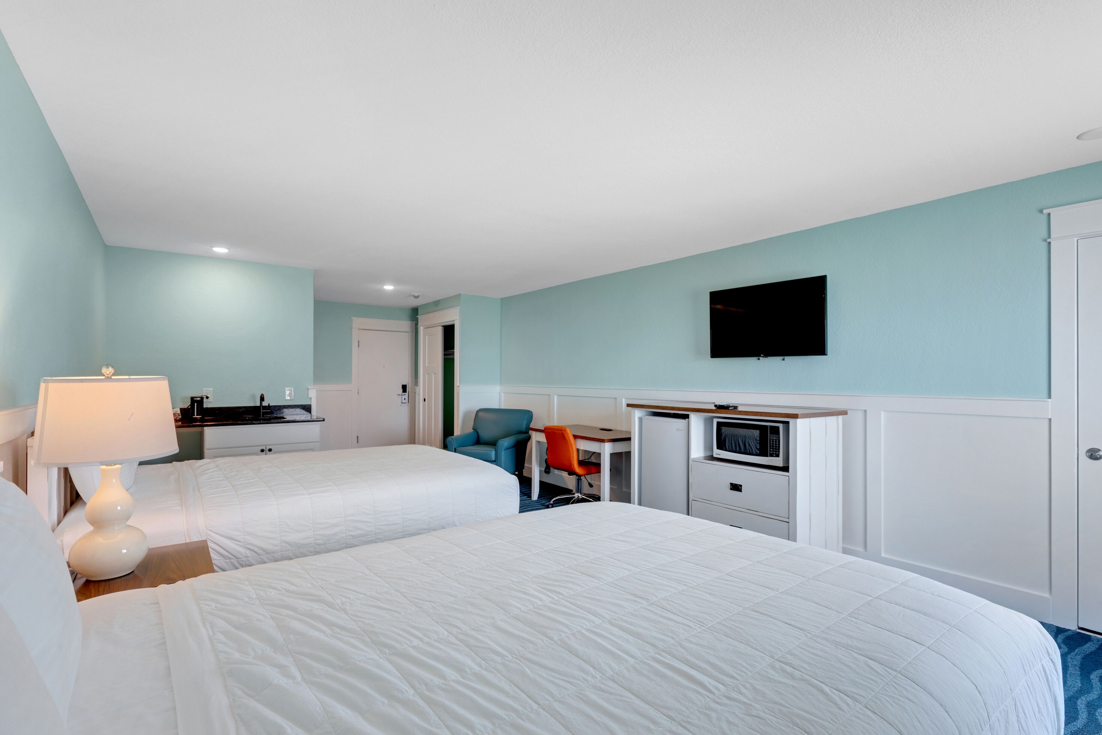A hotel room with two beds, a desk, a chair, a TV, and a kitchenette.