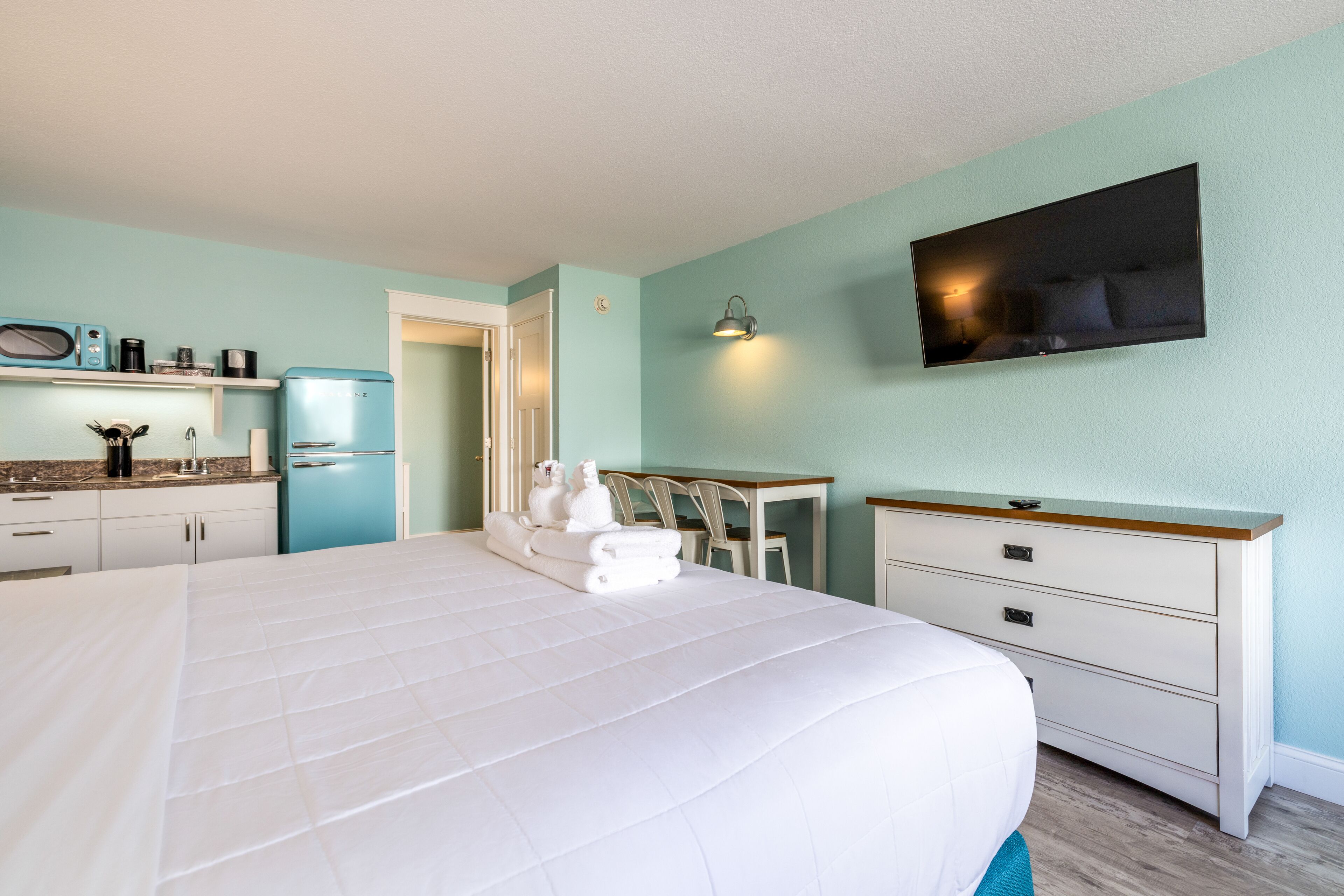 Quarto com uma cama grande, uma escrivaninha, uma cadeira, uma TV na parede e uma cozinha compacta ao fundo.