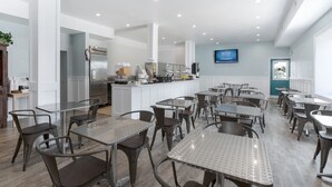 Un interior de restaurante moderno con varias mesas y sillas, una zona de cocina con refrigerador y equipo de cocina, y un televisor montado en la pared.