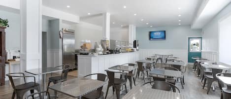 Un intérieur de restaurant moderne avec de nombreuses tables et chaises, un espace cuisine équipé d’un réfrigérateur et d’appareils de cuisson, et un téléviseur fixé au mur.