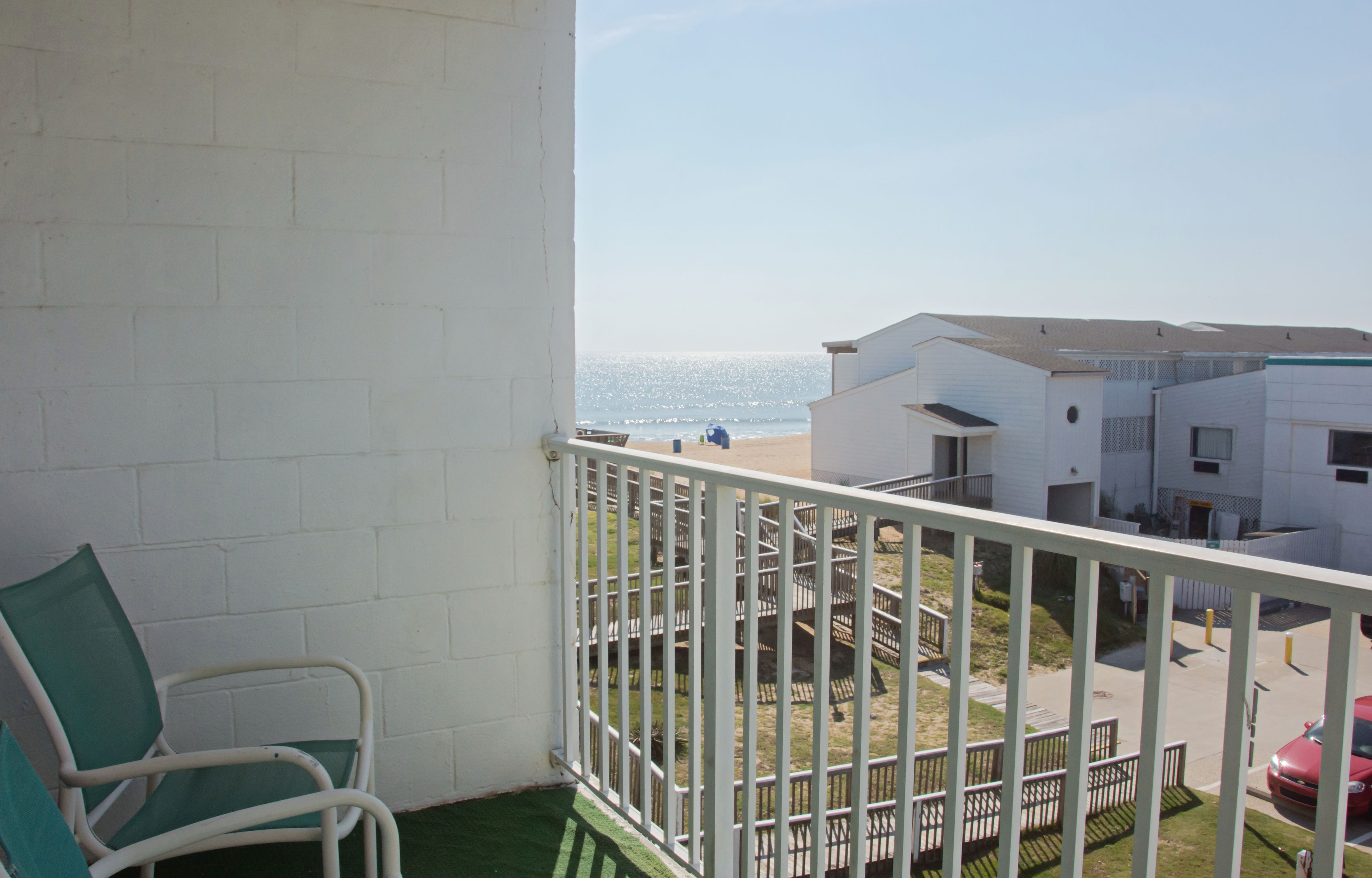 Un balcón con barandilla blanca, una silla verde y vista a la playa y edificios al fondo.
