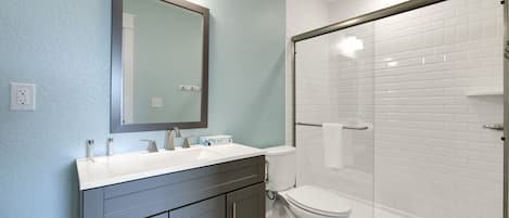 A modern bathroom with a large mirror, a white sink, dark cabinets, a toilet, and a glass-enclosed shower area.