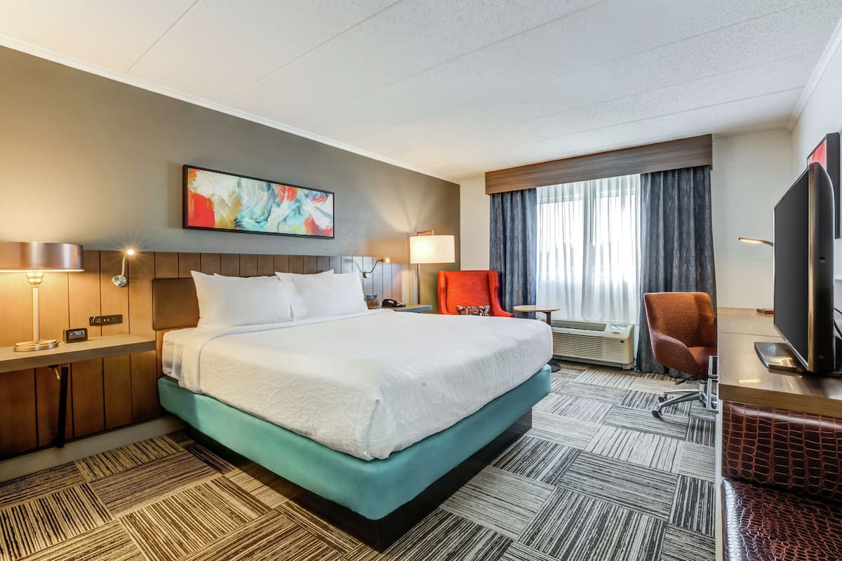A hotel room with a large bed, a desk with a flat-screen TV, a chair, a desk lamp, and a window with curtains.