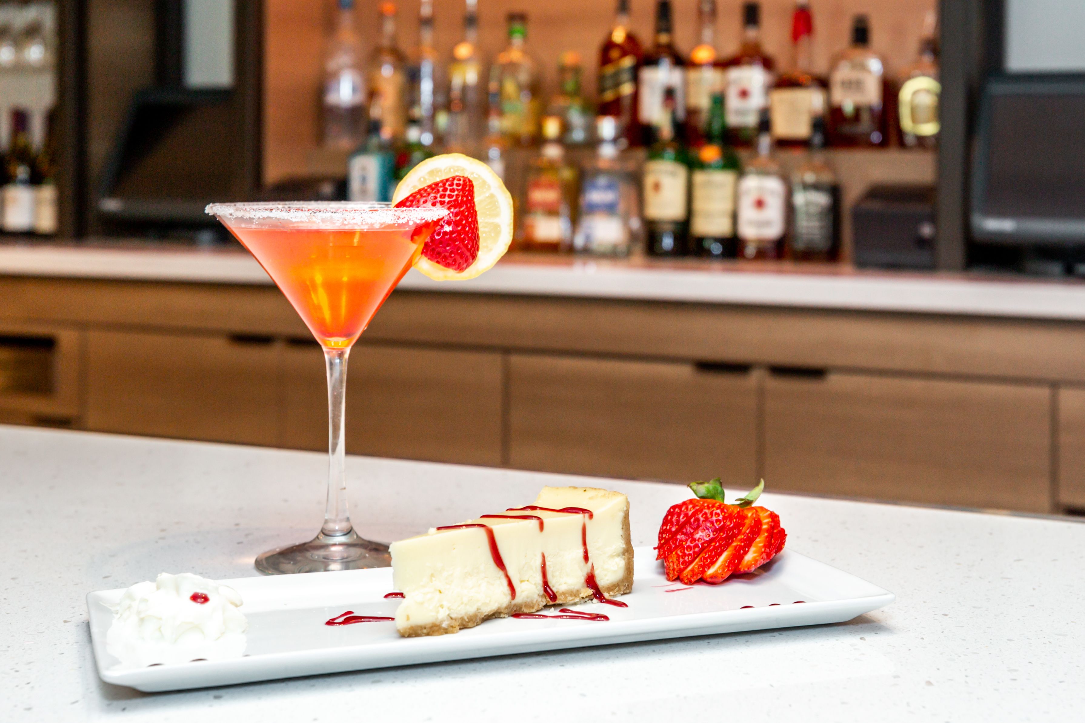 Een cocktail met een aardbeien garnering, een plak cheesecake met aardbeien saus, en een heel aardbei op een wit bord.
