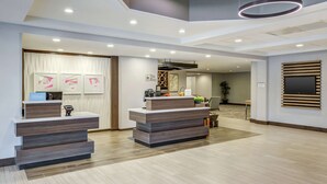 A modern hotel reception area with two reception desks, a large flat-screen TV mounted on the wall, and a ceiling with recessed lighting and circular light fixtures. - Hilton Garden Inn Boston/Waltham (Waltham)