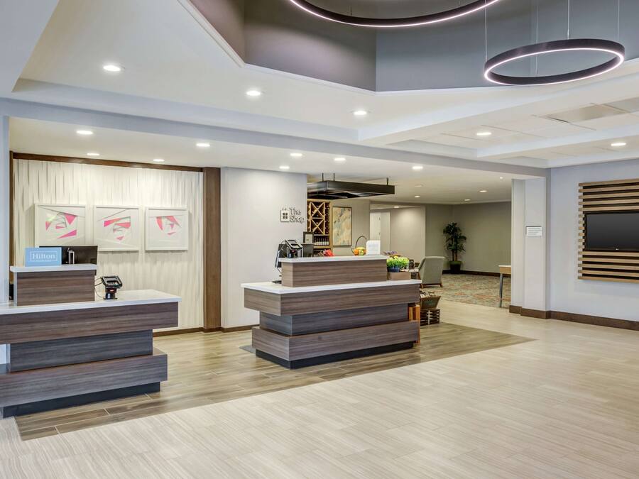 Uma área de recepção de hotel moderna, com duas mesas de atendimento, uma grande TV de tela plana na parede, e um teto com iluminação embutida e luminárias circulares.