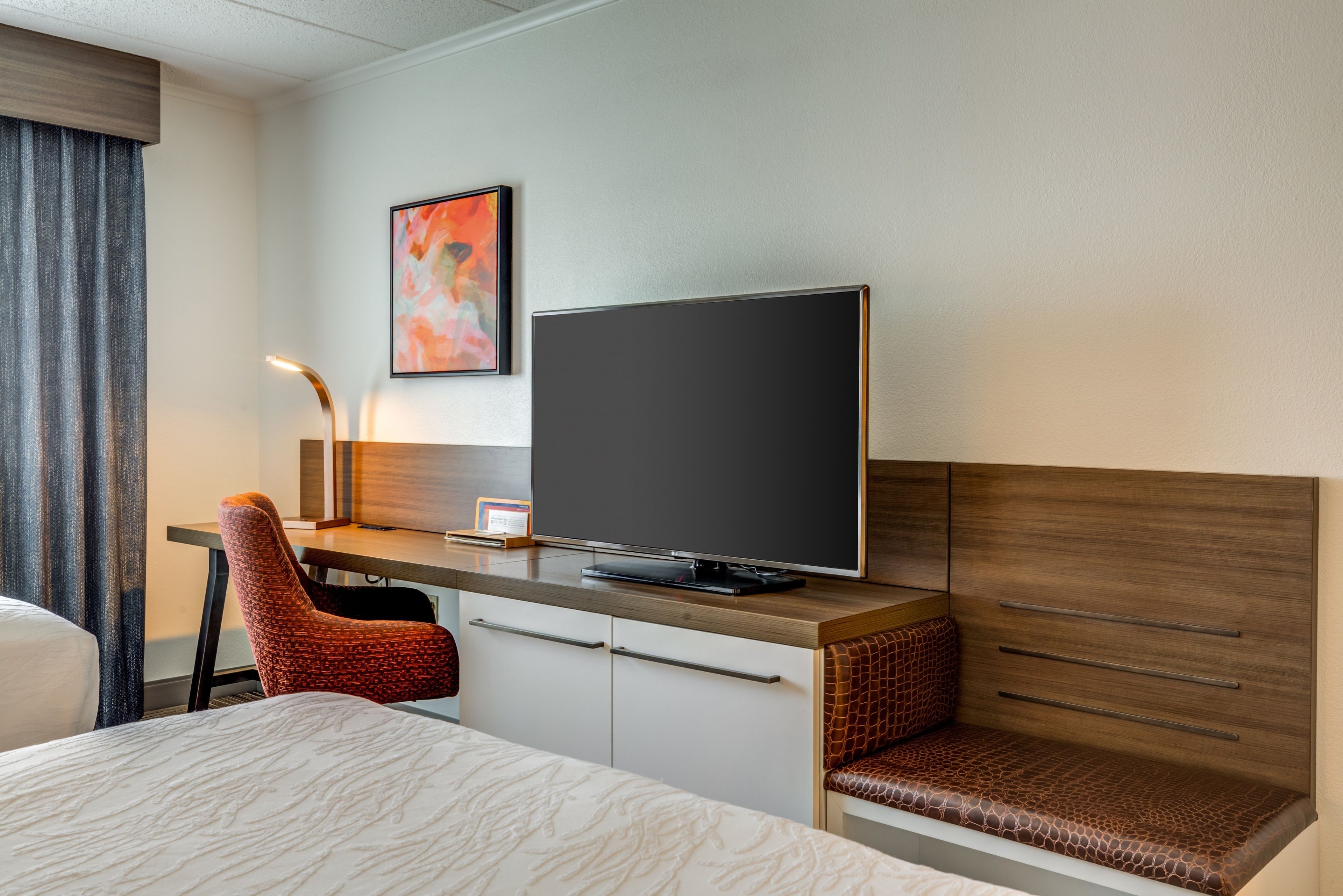 A hotel room with a bed, a desk with a flat-screen TV, a chair, and a wall-mounted shelf.