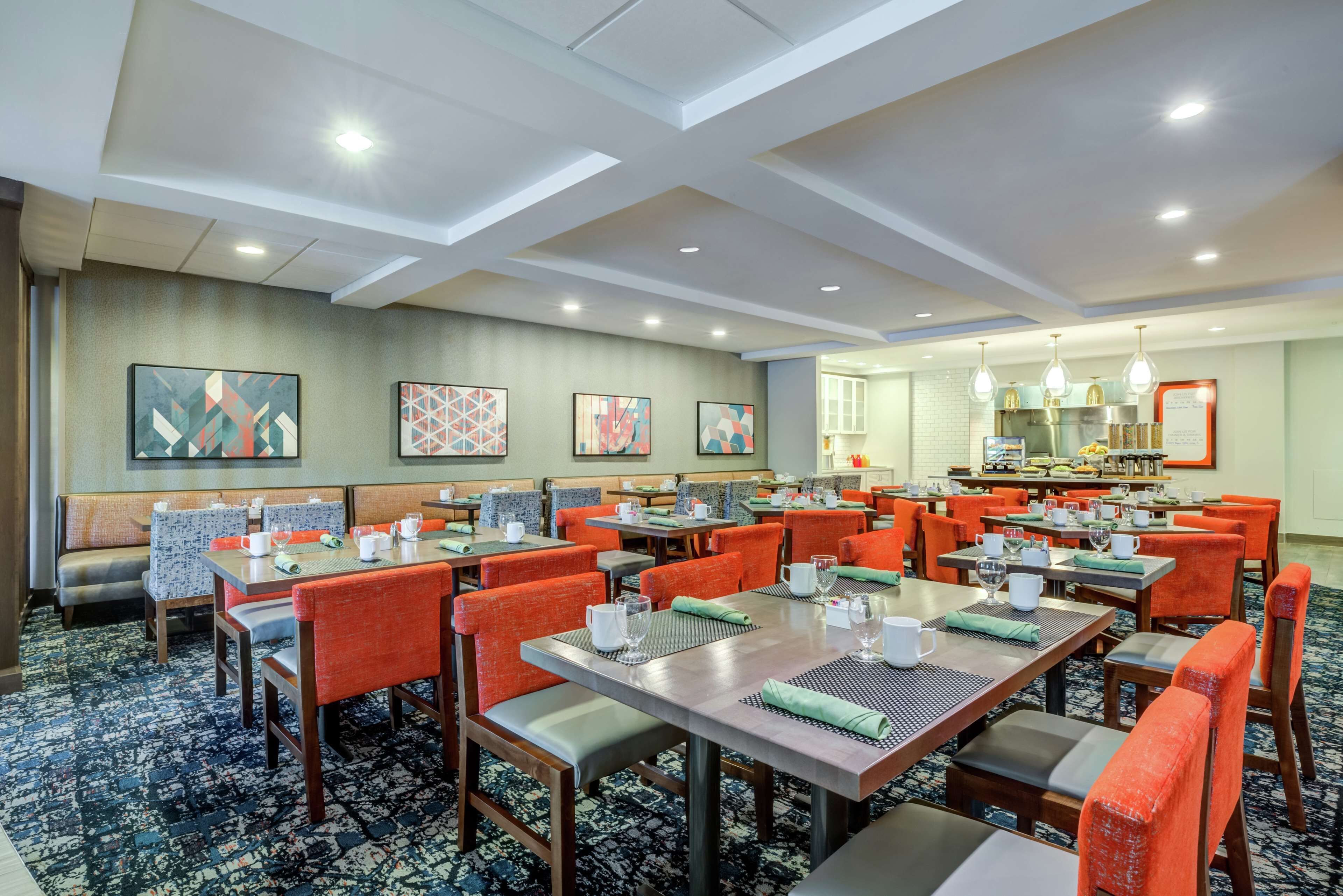 A hotel dining area with multiple tables and chairs, a kitchen in the background, and artwork on the walls.