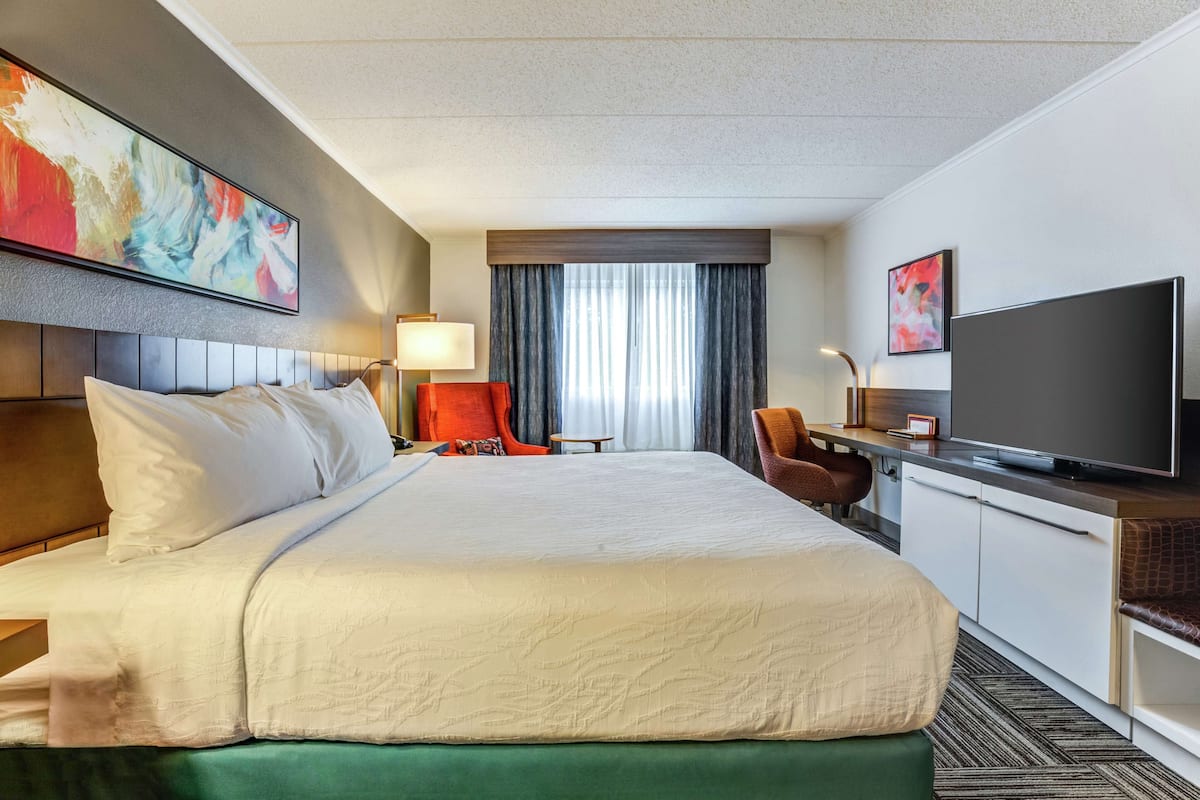 A hotel room with a large bed, a desk with a flat-screen TV, a chair, a lamp, and a window with curtains.
