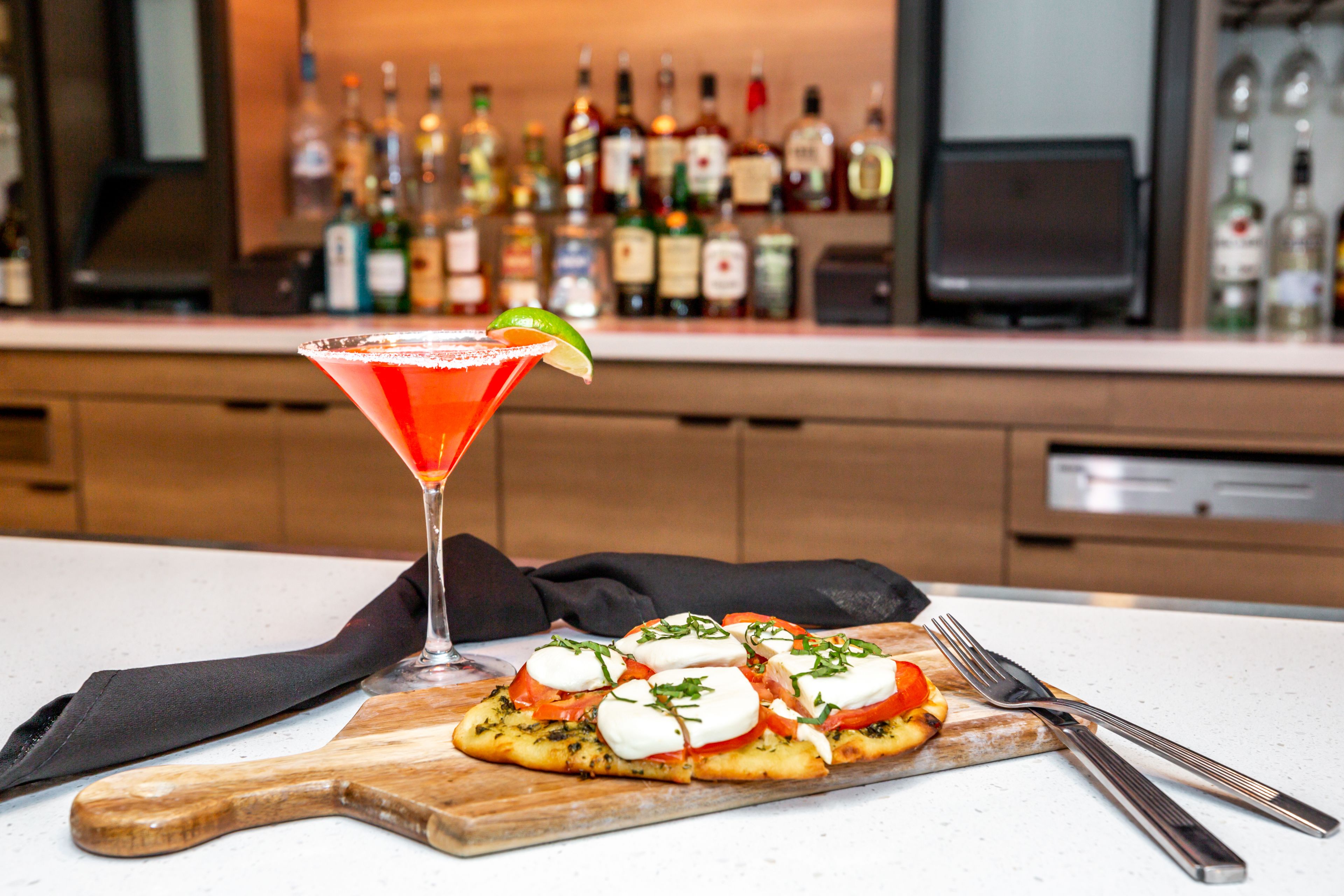 Una copa de martini con un adorno de lima y una tabla de madera con una pizza cubierta de tomates, mozzarella y hierbas.