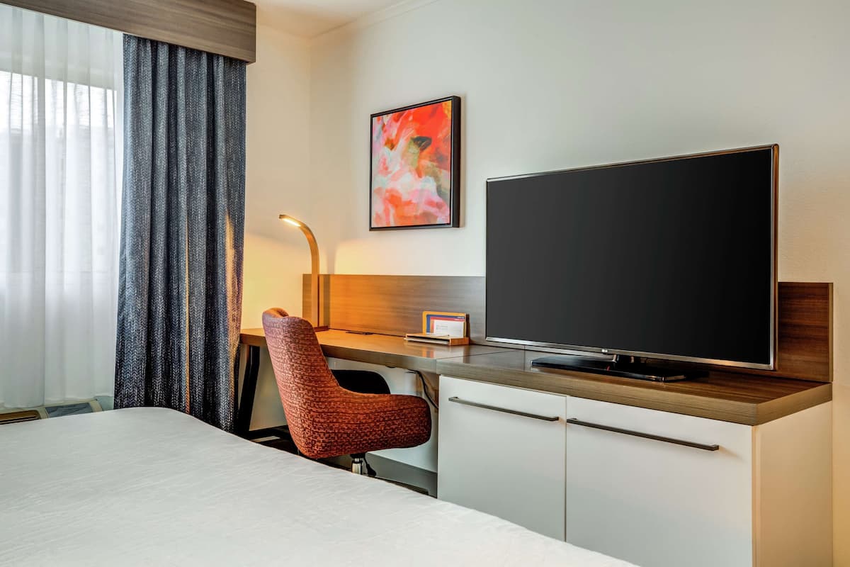 A hotel room with a bed, a desk with a flat-screen TV, a chair, a desk lamp, and a painting on the wall.
