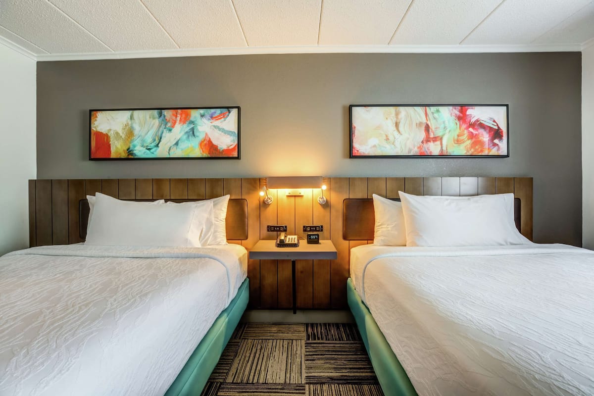 A hotel room with two beds, a wooden headboard, a small table with a phone and a clock, and abstract art on the walls.