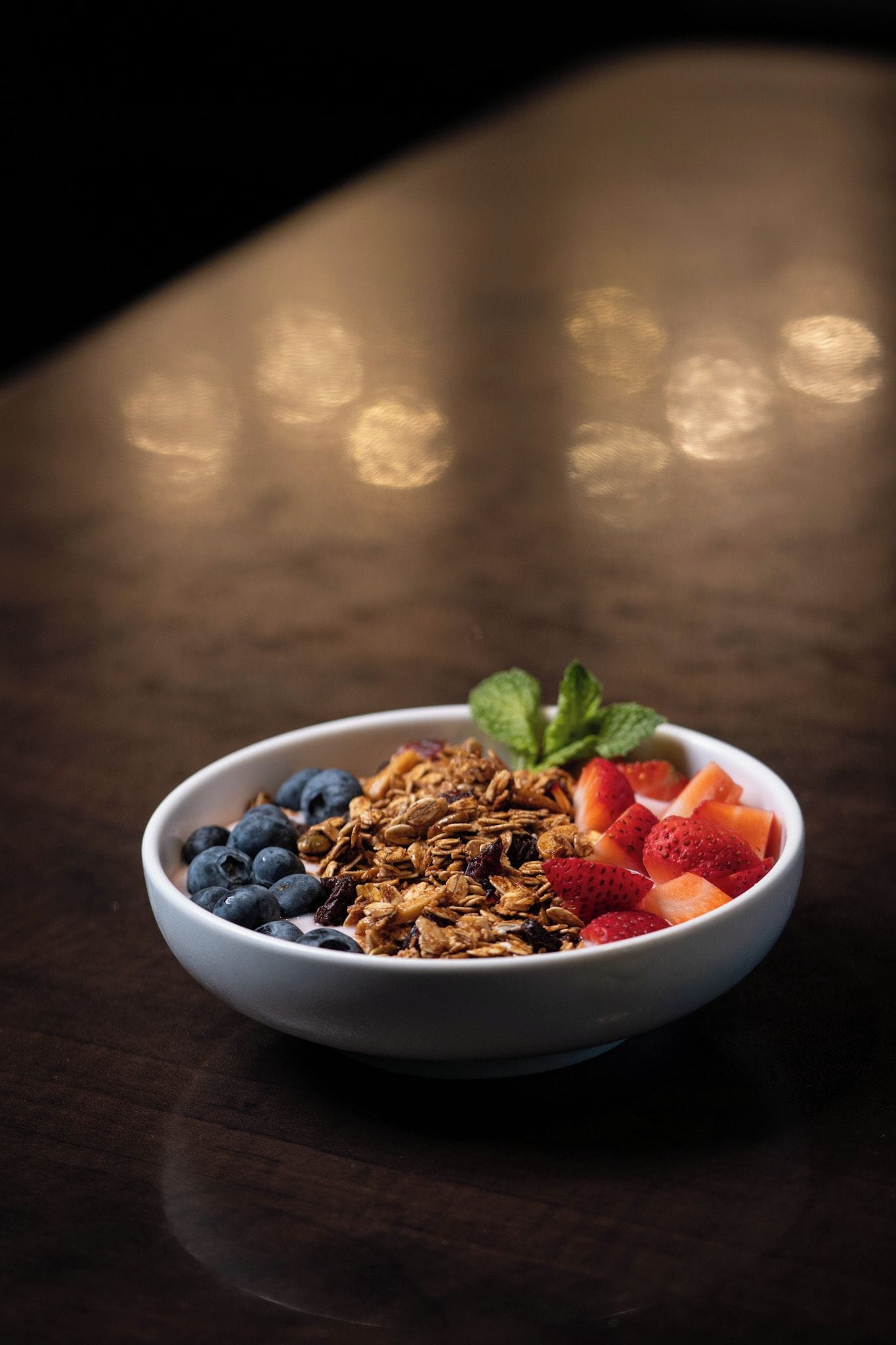 Un bol de granola garni de myrtilles, de fraises et de melon, posé sur une table en bois foncé.