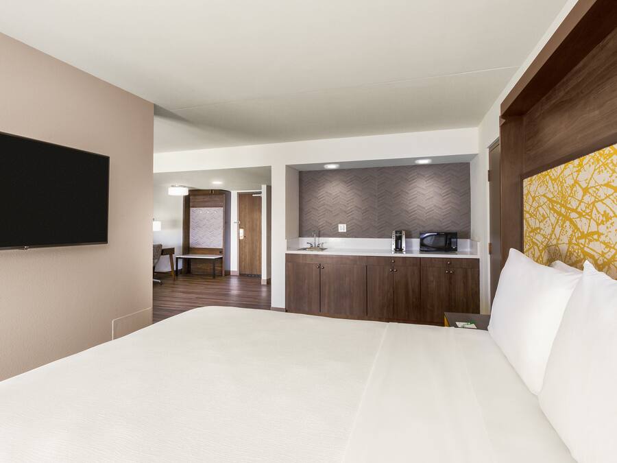 A hotel room with a large bed, a flat-screen TV mounted on the wall, a kitchenette with a microwave, and a herringbone-patterned wall.
