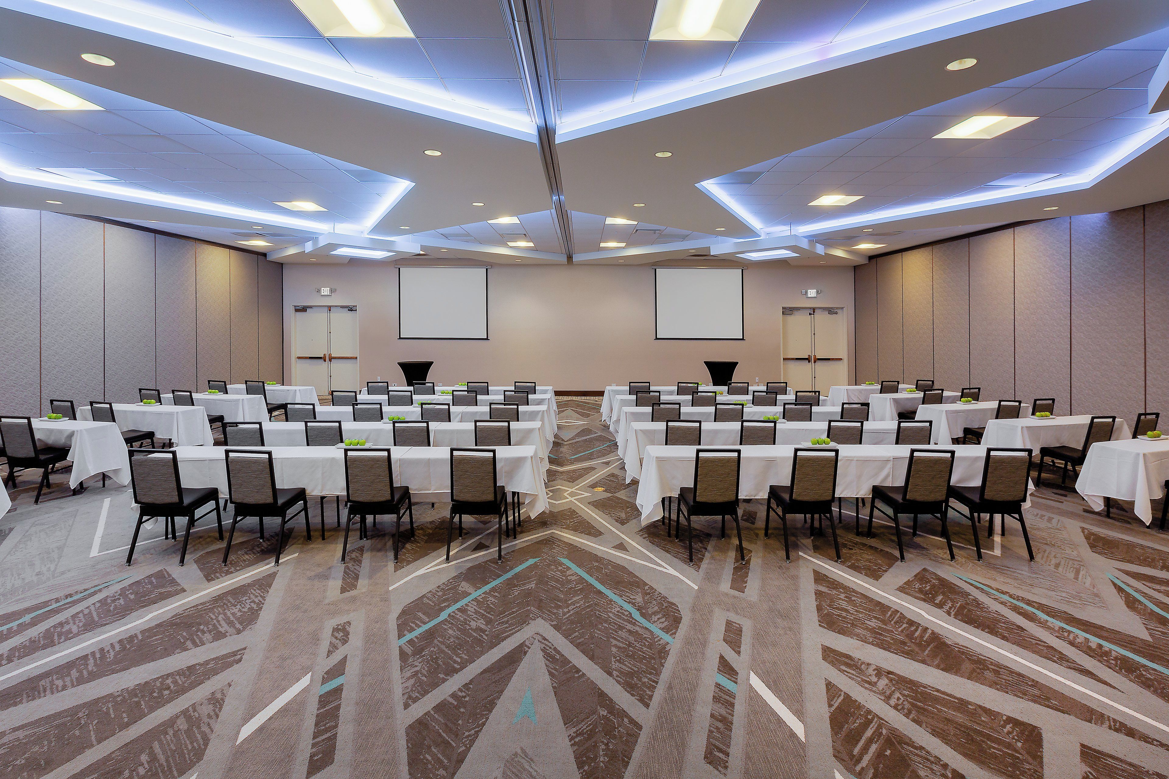 A large conference room with multiple tables and chairs, equipped with a projector screen and a carpeted floor.