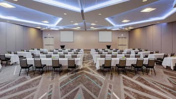 A large conference room with multiple tables and chairs, equipped with a projector screen and a carpeted floor.