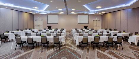 A large conference room with multiple tables and chairs, equipped with a projector screen and a carpeted floor.