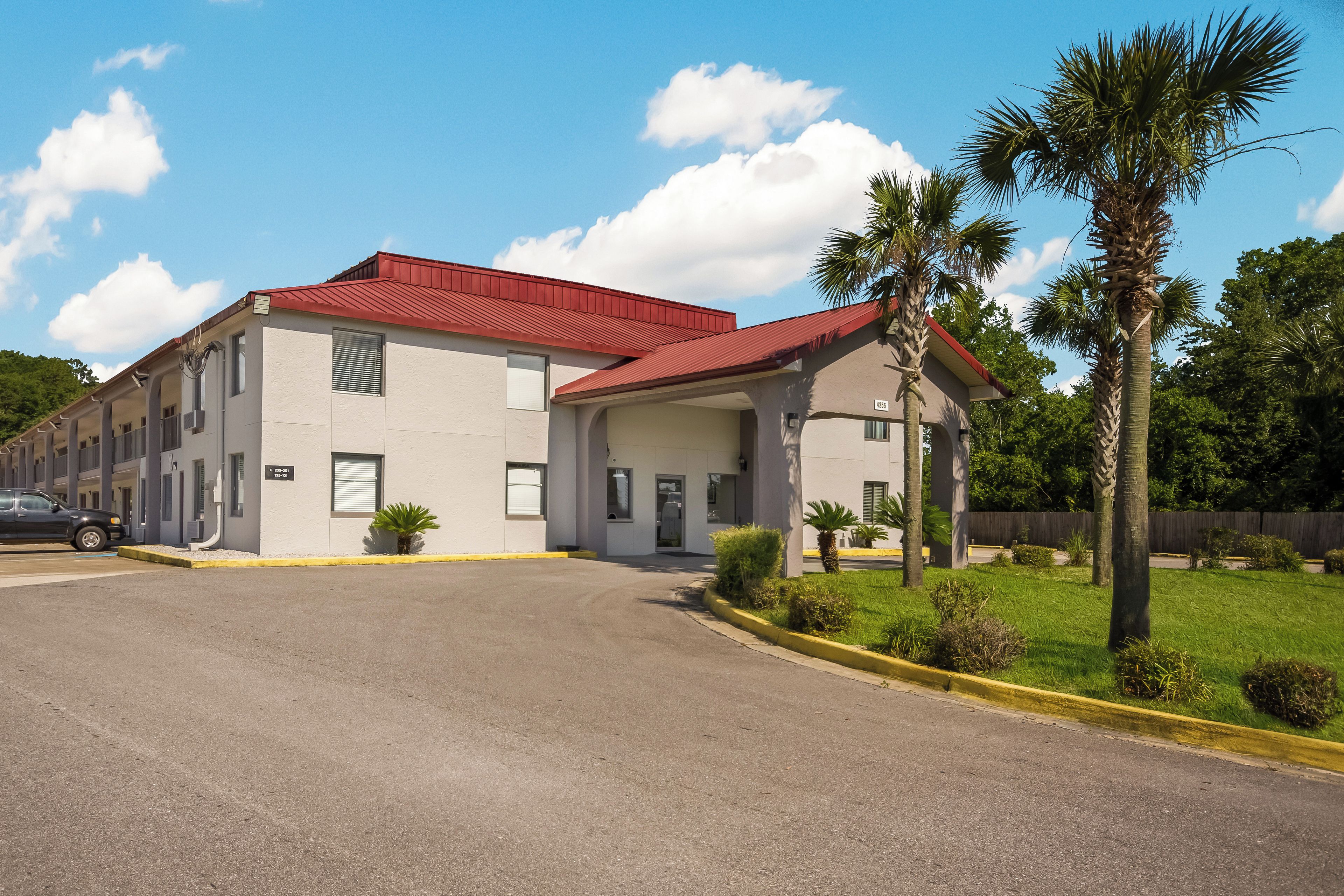 A two-story motel with a red roof, a covered parking area, and a well-maintained lawn with palm trees.