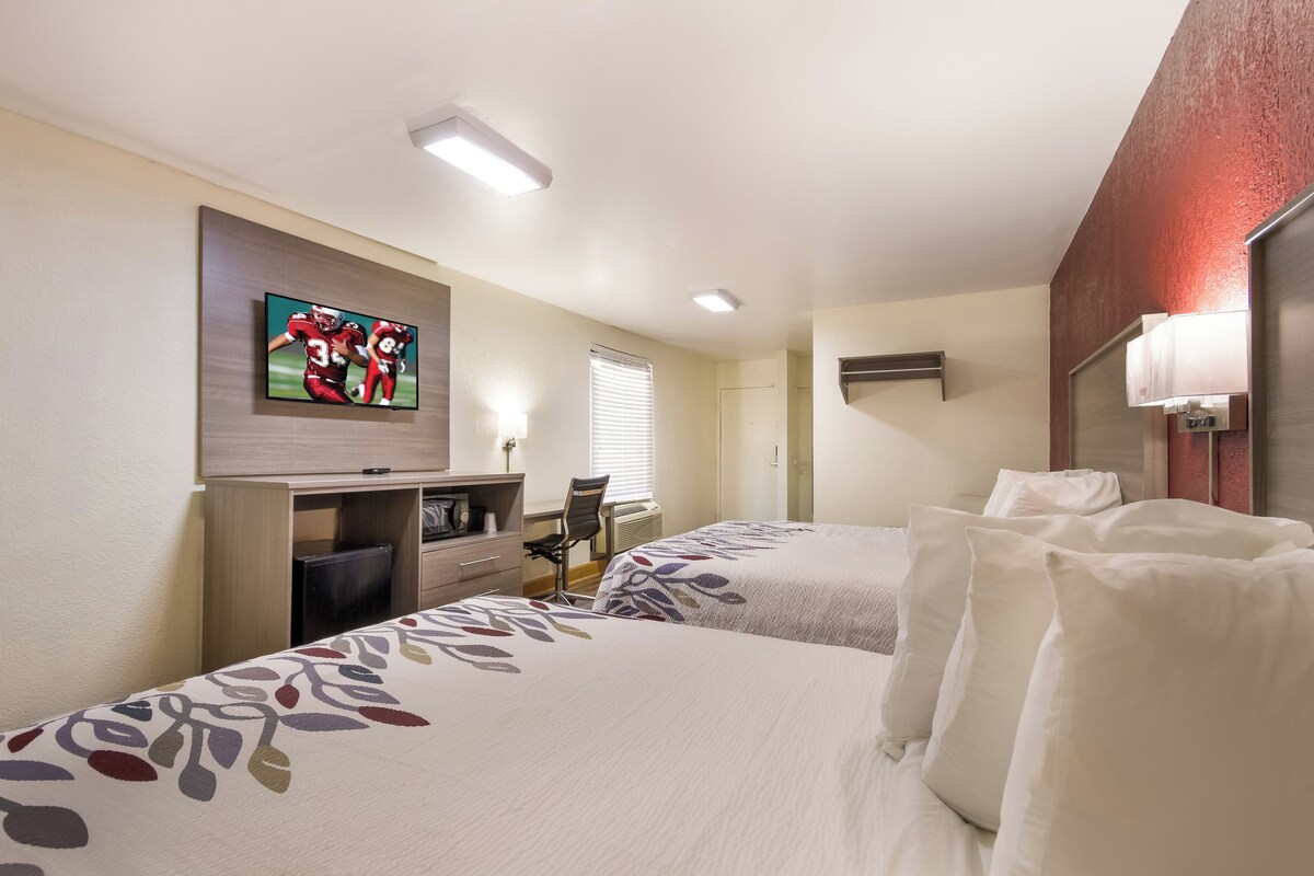 A hotel room with two beds, a desk with a chair, a television mounted on the wall, and a window with blinds.