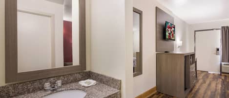 A hotel bathroom with a granite countertop sink, a mirror, and a television mounted on the wall.