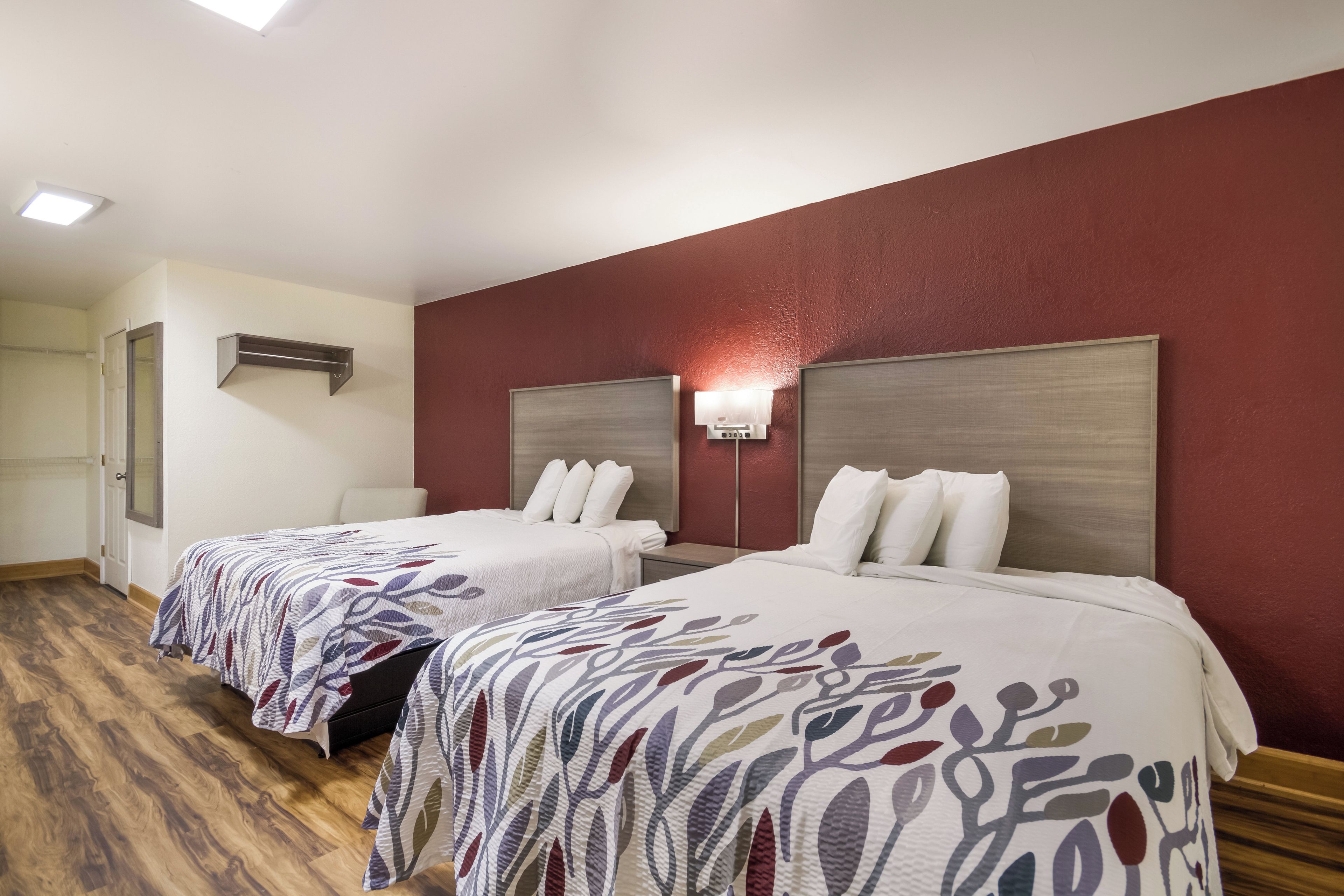 A hotel room with two beds, wooden flooring, a red accent wall, and a ceiling light fixture.